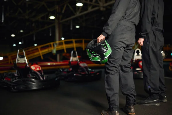Vue recadrée de deux hommes en vêtements de sport debout sur le circuit et tenant des casques près des voitures de karting go — Photo de stock