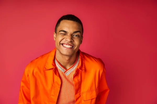 Cheerful african american guy in orange shirt winking at camera on red background — Stock Photo