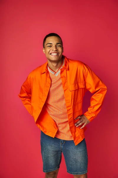 Hombre afroamericano de moda en camisa naranja de pie con las manos en las caderas sobre fondo rojo - foto de stock