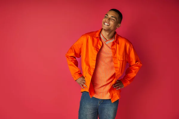 Overjoyed african american in orange shirt, with hands om hips, looking away on red background — Stock Photo