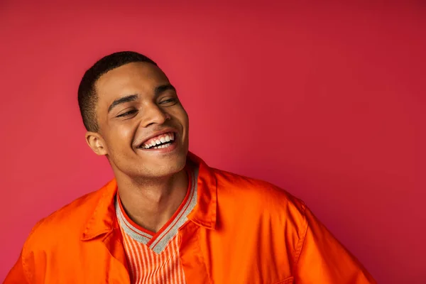 Portrait de mec afro-américain élégant et positif en chemise orange regardant loin sur fond rouge — Photo de stock