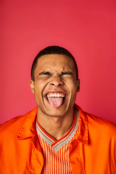 Divertido afroamericano con los ojos cerrados sobresaliendo lengua sobre fondo rojo, camisa naranja, elegante - foto de stock
