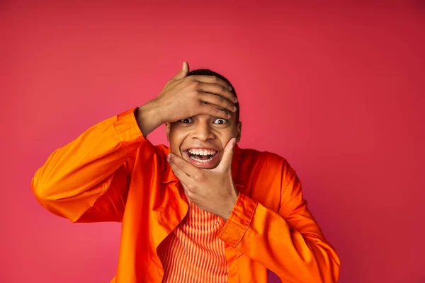 Joyeux afro-américain mec toucher le visage et regarder la caméra, fond rouge, chemise orange — Photo de stock
