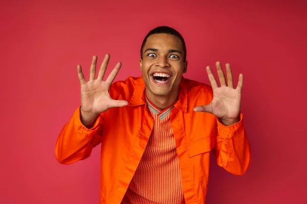 Playful african american man with crazy face expression showing scary gesture on red, orange shirt — Stock Photo