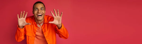 Funny african american guy showing scary gesture and grimacing on red, orange shirt, banner — Stock Photo