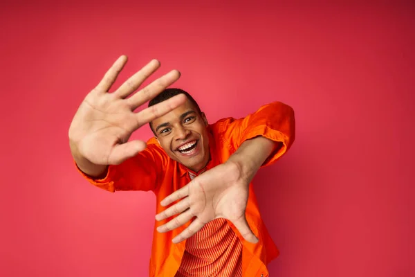 Alegre afroamericano hombre con las manos extendidas mirando a la cámara en rojo, camisa naranja, de moda - foto de stock