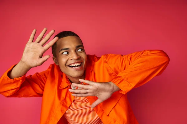 Animado e engraçado homem americano africano gesticulando e olhando para longe em vermelho, camisa laranja, na moda — Fotografia de Stock