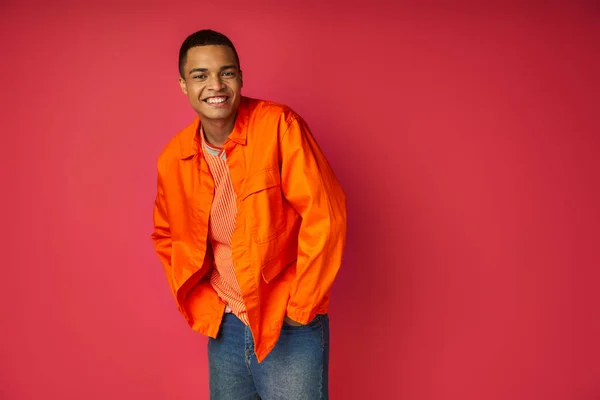Модный африканский американец с руками в кармане, улыбающийся в камеру на красном фоне — стоковое фото