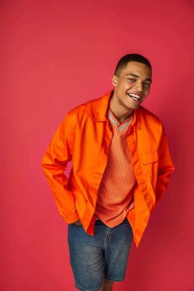 Joyful african american guy in orange shirt, with hands in pockets, smiling at camera on red — Stock Photo