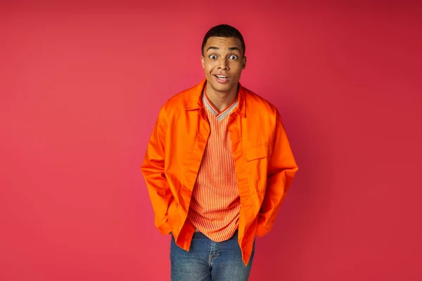 Surpreso afro-americano homem com as mãos no bolso, em camisa laranja olhando para a câmera em vermelho — Fotografia de Stock