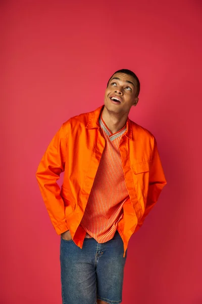 Positivo y curioso afroamericano en camisa naranja mirando hacia arriba sobre fondo rojo, manos en bolsillos - foto de stock