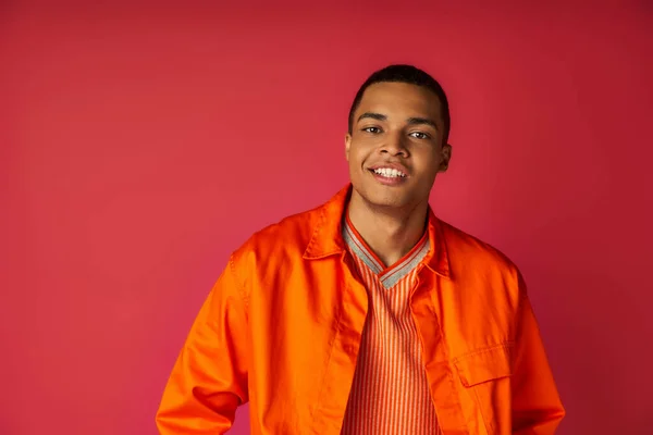 Chico afroamericano guapo en camisa naranja de moda, con sonrisa radiante mirando a la cámara en rojo - foto de stock