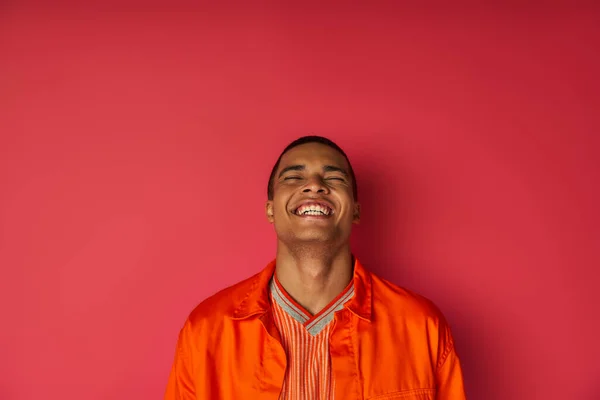 Homme afro-américain ravi en chemise orange riant les yeux fermés sur le rouge, excitation — Photo de stock