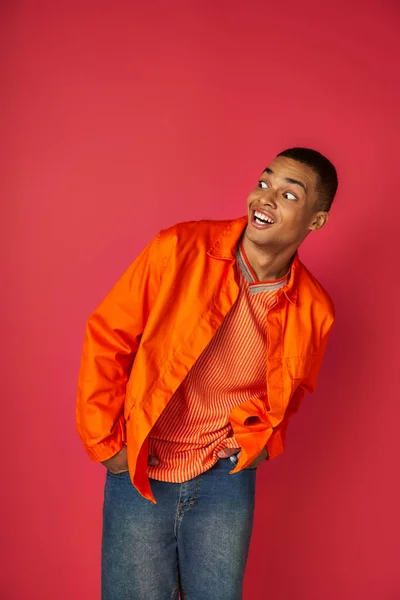 Homem afro-americano surpreso sorrindo e olhando para o vermelho, mãos nos bolsos, camisa laranja — Fotografia de Stock