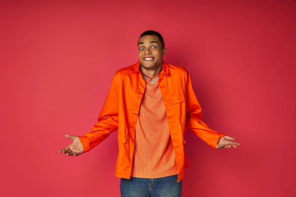 Зачарований афроамериканський чоловік в помаранчевій сорочці, що показує жест згинання на червоному тлі — стокове фото