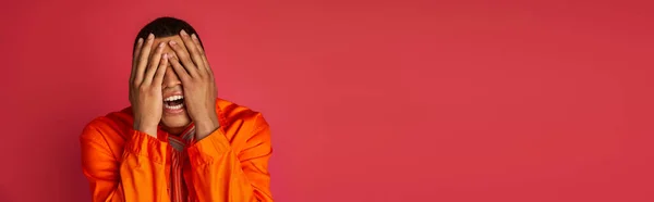 Homme afro-américain bouleversé en chemise orange obscurcissant visage avec les mains sur rouge, bannière, espace de copie — Photo de stock