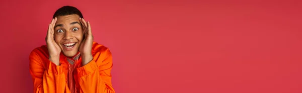 Радостный африканский американец, руки возле лица, смотрящий на камеру на красном, оранжевая рубашка, баннер, копировальное пространство — стоковое фото