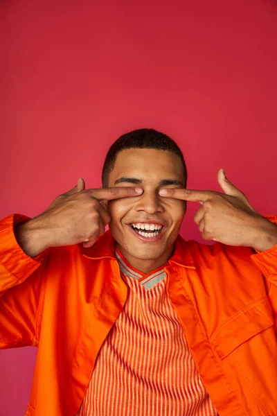 Fröhlicher afrikanisch-amerikanischer Typ, der die Augen mit Fingern verdeckt, orangefarbenes Hemd, roter Hintergrund — Stockfoto