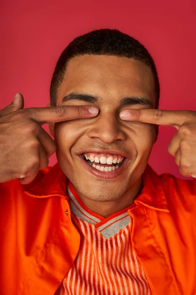 Hombre afroamericano divertido oscurecer los ojos con los dedos, camisa naranja, fondo rojo - foto de stock