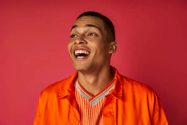 Homme afro-américain étonné en chemise orange regardant loin et riant sur fond rouge — Photo de stock