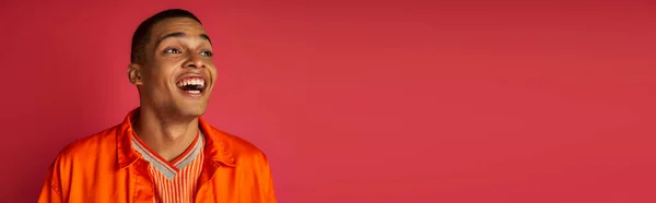 Asombrado hombre afroamericano, riendo, mirando hacia otro lado, camisa naranja, fondo rojo, pancarta - foto de stock