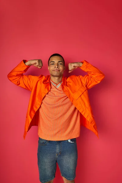 Fiducioso uomo africano americano in camicia arancione dimostrando forza su sfondo rosso — Foto stock