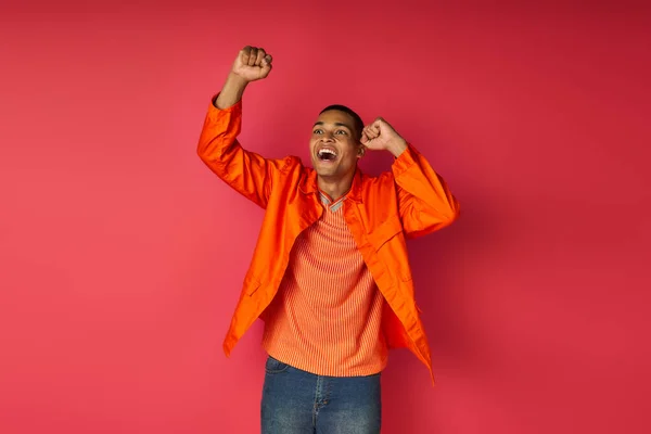 Aufgeregter afrikanisch-amerikanischer Typ in orangefarbenem Hemd zeigt Erfolgsgeste und schreit auf rotem Hintergrund — Stockfoto