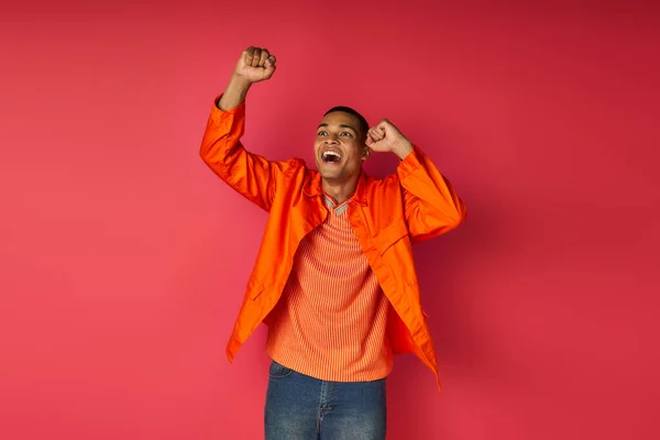 Excited african american guy in orange shirt showing success gesture and shouting on red background — Stock Photo