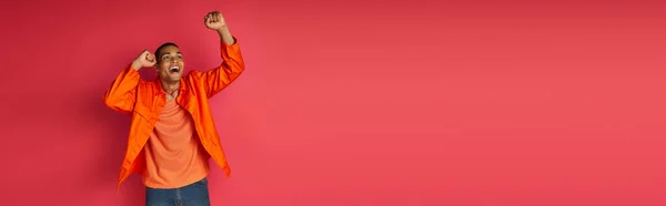 Веселий афроамериканський чоловік в помаранчевій сорочці кричить і показує жест перемоги на червоному тлі — стокове фото