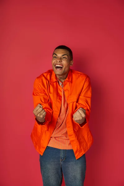 Рад, взволнован африканский американец кричать и показывать победу жест, оранжевая рубашка, красный фон — стоковое фото