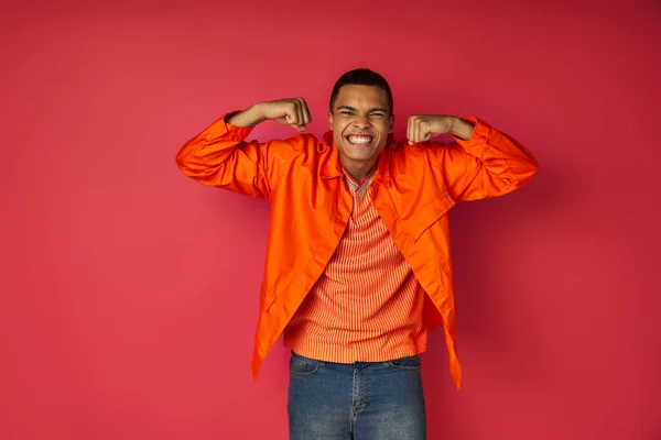Homme afro-américain expressif en chemise orange démontrant la force et regardant la caméra sur rouge — Photo de stock