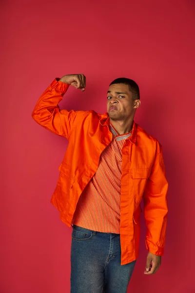 Haughty african american guy demonstrating strength, looking at camera, orange shirt, red background — Stock Photo