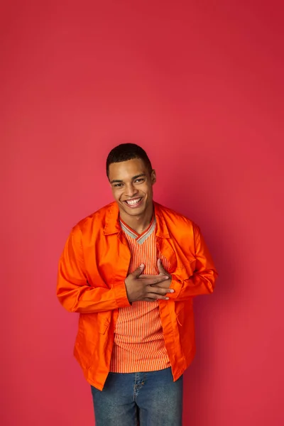 Щасливий афроамериканський чоловік торкається грудей, посміхається на камеру на червоному, вдячний жест, помаранчева сорочка — стокове фото
