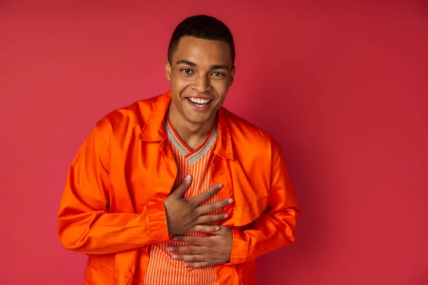 Joyful african american guy in orange shirt showing grateful gesture and touching chest on red — Stock Photo