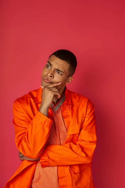 Profundamente en el pensamiento hombre afroamericano en camisa naranja tocando el pecho y mirando hacia otro lado en rojo - foto de stock