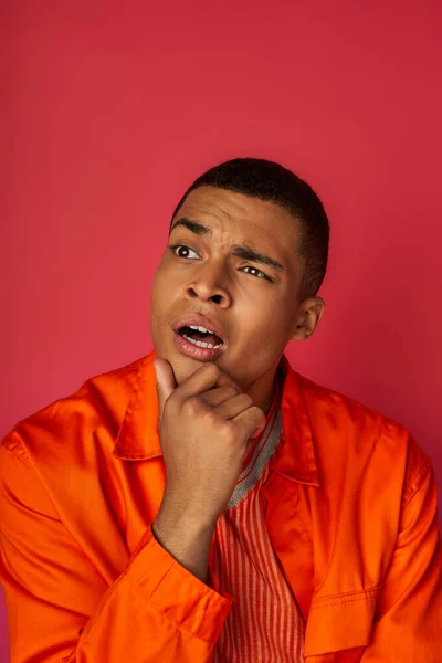 Hombre afroamericano pensativo en camisa naranja tocando el pecho, pensando, mirando hacia otro lado en rojo - foto de stock