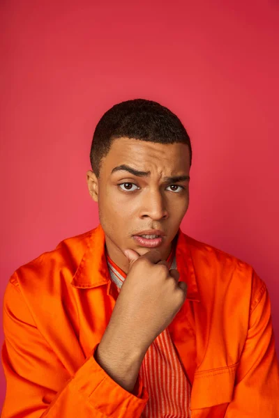 Homme afro-américain réfléchi et inquiet en chemise orange touchant la poitrine, regardant la caméra sur le rouge — Photo de stock