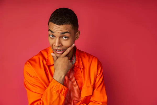 Entmutigte, nachdenkliche afrikanisch-amerikanische Mann in orangefarbenem Hemd berührt Kinn, Blick in die Kamera auf rot — Stockfoto