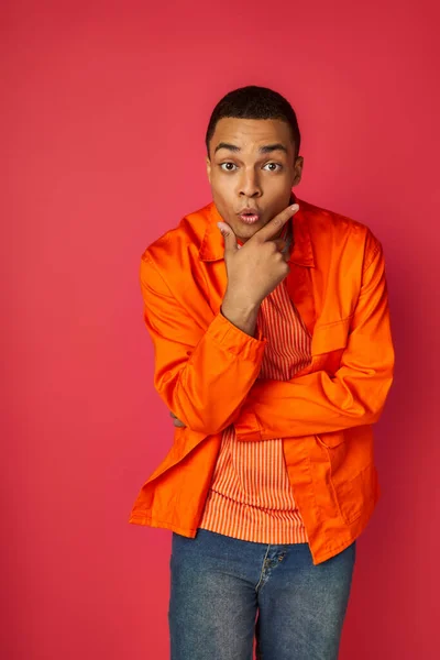 Homme afro-américain excité en chemise orange, avec la main près du visage regardant la caméra sur le rouge — Photo de stock