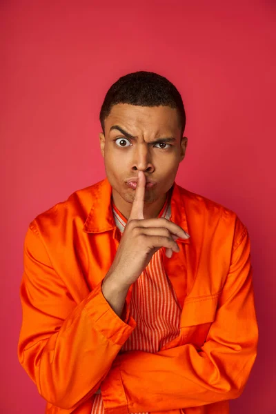 Strenger afrikanisch-amerikanischer Mann in orangefarbenem Hemd blickt in die Kamera und zeigt Schweigegegeste auf Rot — Stockfoto