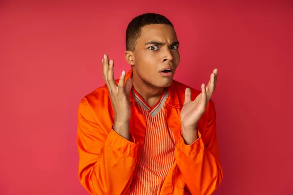 Mécontent, choqué afro-américain en chemise orange geste, en regardant la caméra sur fond rouge — Photo de stock