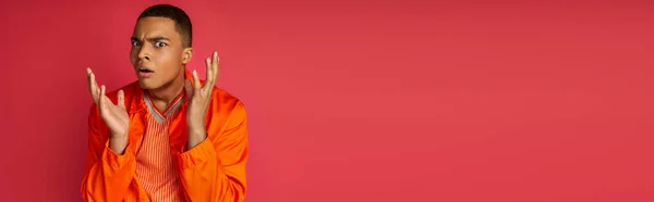 Scioccato afroamericano in camicia arancione gesticolando e guardando la fotocamera su rosso, banner, spazio copia — Foto stock