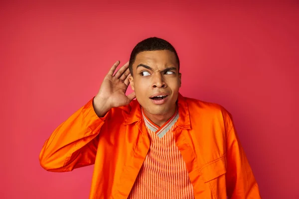 Curioso uomo afroamericano con mano vicino orecchio origliare su sfondo rosso — Foto stock