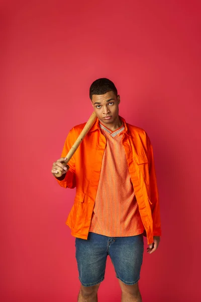 Confiado hombre afroamericano en camisa naranja sosteniendo bate de béisbol y mirando a la cámara en rojo - foto de stock