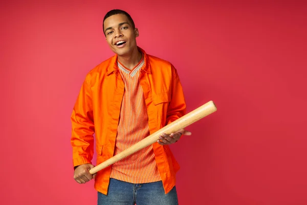 Sorridente, scettico afroamericano con mazza da baseball che guarda la macchina fotografica su sfondo rosso — Foto stock
