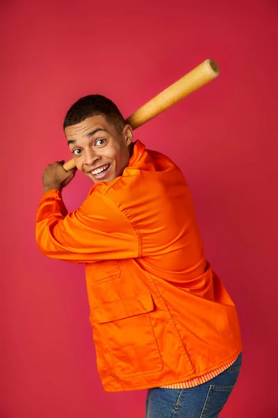 Энергичный африканский американец в оранжевой рубашке играет в бейсбол на красном фоне — стоковое фото