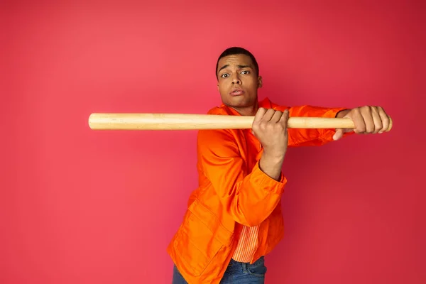 Concentrated african american man in orange shirt holding baseball bat and looking at camera on red — Stock Photo