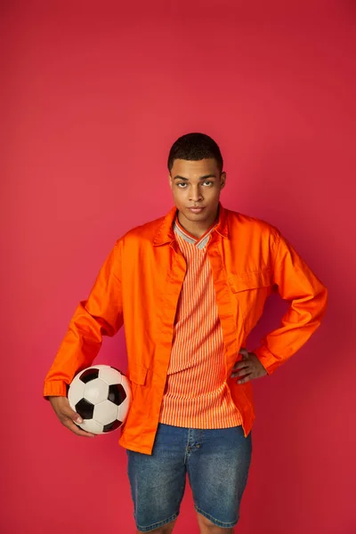 Confident african american in orange shirt standing with soccer ball and hand on hip on red — Stock Photo