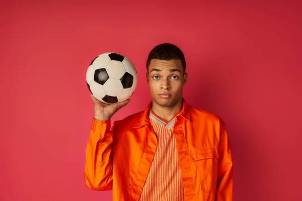 Youthful african american guy in orange shirt holding soccer ball and looking at camera on red — Stock Photo