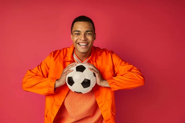 Joyful african american man in orange shirt holding soccer ball and smiling at camera on red — Stock Photo